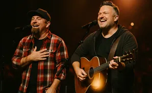 Two men singing and playing guitar on stage with microphones and warm lighting.