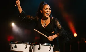 A woman with a peace sign necklace plays drums on stage, smiling and holding drumsticks.