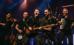 A band performs on stage. From left to right, a man plays the violin, another plays bagpipes and guitar, a third plays guitar, and the fourth plays the …