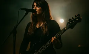 A woman sings into a microphone while playing a black bass guitar on stage, with a bright light behind her.