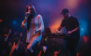 A female singer performs into a microphone while a DJ plays music behind her.