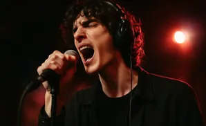 A man with curly hair sings passionately into a microphone while wearing headphones, lit by red stage lights.