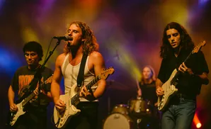 A band performs on stage, with three guitarists and a drummer visible.