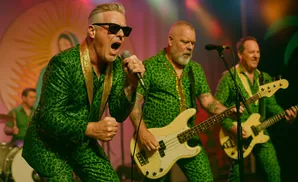 A band performs on stage, wearing matching green leopard print suits with gold trim. The lead singer, in sunglasses, sings into a microphone.
