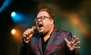 A man wearing glasses and a floral suit sings into a microphone on stage with colorful lights in the background.