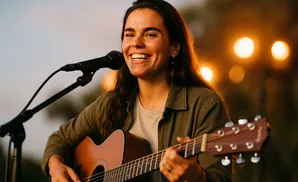 A woman smiles while playing an acoustic guitar and singing into a microphone.