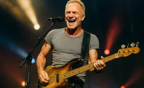 Sting sings and plays bass guitar on stage with bright stage lights.