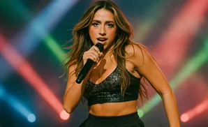 Singer performing on stage, holding microphone, wearing a sequined top, with colorful lights in the background.