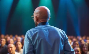 Bald man in a blue shirt giving a speech to a blurred audience under stage lights.