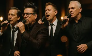 Four men in suits sing into microphones in a dimly lit setting.