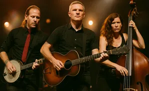 A band of three musicians performing on stage. A man on the left plays a banjo, a man in the center plays guitar, and a woman on the right plays a doubl…