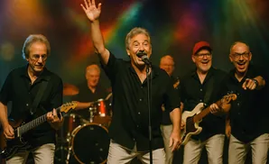 A band of five men on stage performing with guitars and drums. The lead singer waves at the audience.
