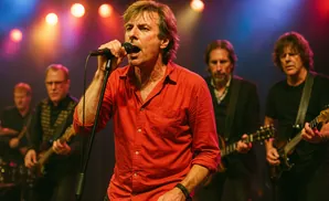 A rock band performs on stage, with the lead singer in a red shirt in focus, singing into a microphone.