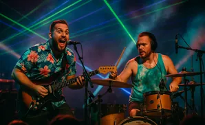 A guitarist sings into a microphone while playing his guitar, with a drummer behind him playing a drum set.
