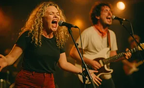 A female singer with blonde curly hair belts into a microphone, with a male guitarist behind her.