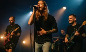 A rock band performs on stage with a singer in the foreground holding a microphone and a bassist to his right playing a teal bass guitar.