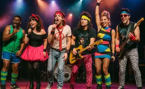 A diverse band of six singers and musicians dressed in vibrant 1980s attire perform on stage.