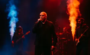 Singer with microphone on stage with band, lit by red and blue smoke effects.