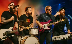 A band performs on stage. A singer with glasses and a beard sings into a microphone, with a guitarist, drummer, and keyboardist behind him.