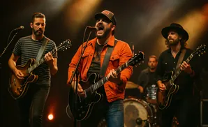 Band performing live on stage, with singers and guitarists in the foreground, and a drummer in the background.