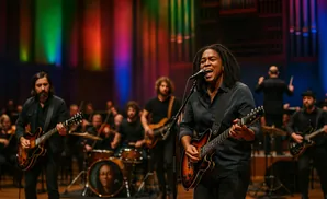 A Black woman sings and plays guitar on stage, with an orchestra and other musicians behind her.