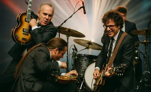 Band in suits playing guitars and drums on stage with a colorful backdrop.