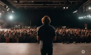 A person stands on a stage facing a large, cheering crowd under bright stage lights.