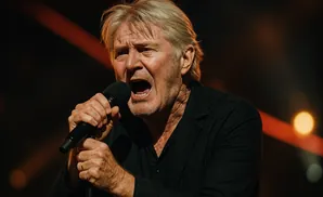 A man with gray hair sings into a microphone, his mouth open in a powerful expression.