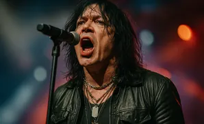 A rock singer in a black leather jacket sings passionately into a microphone, with lights in the background.
