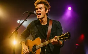 A man with blond hair sings into a microphone while playing an acoustic guitar on stage.
