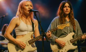 Two women playing guitars on stage under warm lighting.