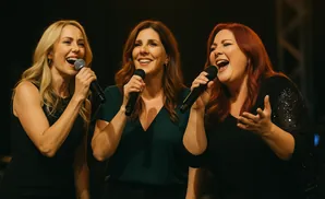 Three women singing into microphones on a stage.