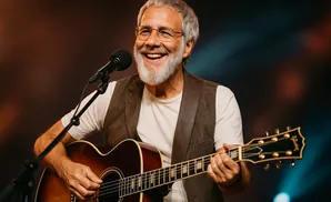 An older man with gray hair and a beard plays an acoustic guitar and sings into a microphone.