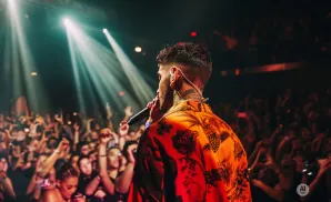 A performer sings into a microphone on a stage, illuminated by spotlights, with a cheering crowd visible in the background.