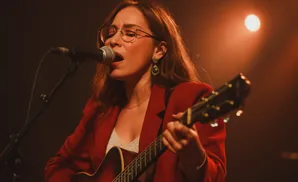 Woman in a red jacket singing into a microphone while playing an acoustic guitar.