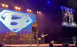 A Black male singer in a Superman-style shirt performs on stage with a band, with a large 