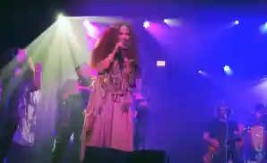 Singer with long, dark, voluminous hair sings into a microphone on a stage with a band, illuminated by green and purple lights.