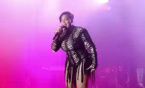 A woman in a revealing black dress sings into a microphone on a stage lit with pink and purple lights.