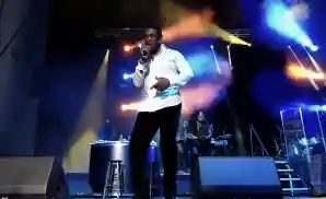 A man in a white shirt and black pants sings into a microphone on a stage with colorful lights.