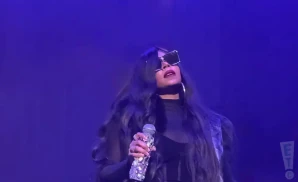 Singer with long dark hair, large sunglasses, and a bedazzled microphone performs on a purple stage.