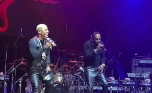 Two men in leather pants sing into microphones on a dimly lit stage with a drum set and keyboards.