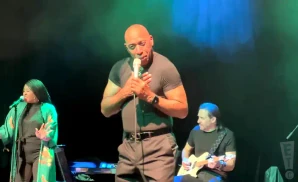 A man in a black t-shirt sings into a microphone on stage, with two musicians beside him.