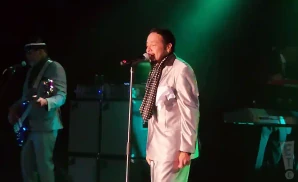 A singer in a white suit and polka dot scarf performs on stage, while a bassist in a light blue suit plays on the left.