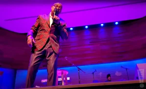 A Black man in a patterned suit sings into a microphone on a stage with purple lighting.