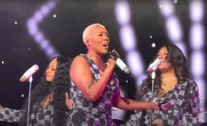 Singer with a blonde buzz cut and two backing singers perform on stage.