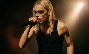 A male singer with long, blonde hair and heavy eye makeup holds a microphone to his lips, wearing a black tank top.
