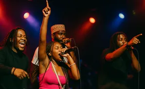 A woman in a pink tank top sings into a microphone, while three men with microphones perform on a dimly lit stage.