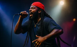 A Black man with dreadlocks and a red cap sings into a microphone, wearing a dark shirt and gold necklaces.