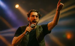 A man in glasses and a vest sings into a microphone with his arm raised, pointing his finger upwards.