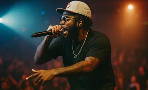 A male rapper in a black t-shirt and baseball cap performs into a microphone on stage, lit by blue and orange spotlights.